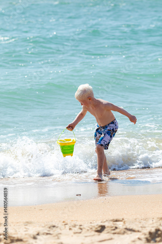 Obraz na płótnie Happy child boy playing with the sand on the beach, pouring water from plastic bucket on the sea beach, building sand castle, enjoying summer. Advertising for tour operators, hotels, airlines. w salonie