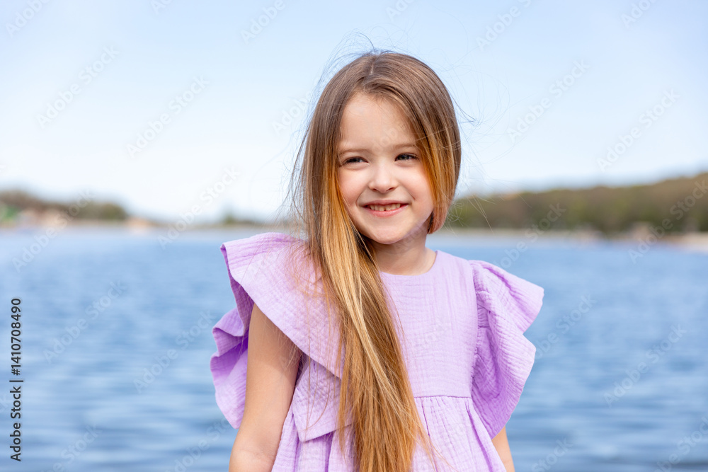 Happy little girl with long blonde hair, in lilac summer dress, sitting on the beach with blue water background. Children's Day. Vacation concept. Space for text. Advertising for tour operators