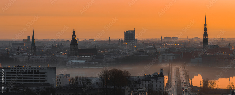 A warm sunrise illuminates Riga's old town, featuring St. Peter's Church, Riga Cathedral, a bridge over the Daugava River, and light morning mist.