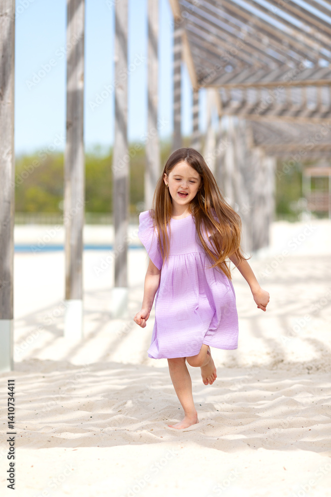 Carefree summer. Little girl in lilac dress has fun and running barefoot on sandy beach under wooden gazebo on sunny day. Joyful childhood adventures and tropical vacation concept. Space for text.