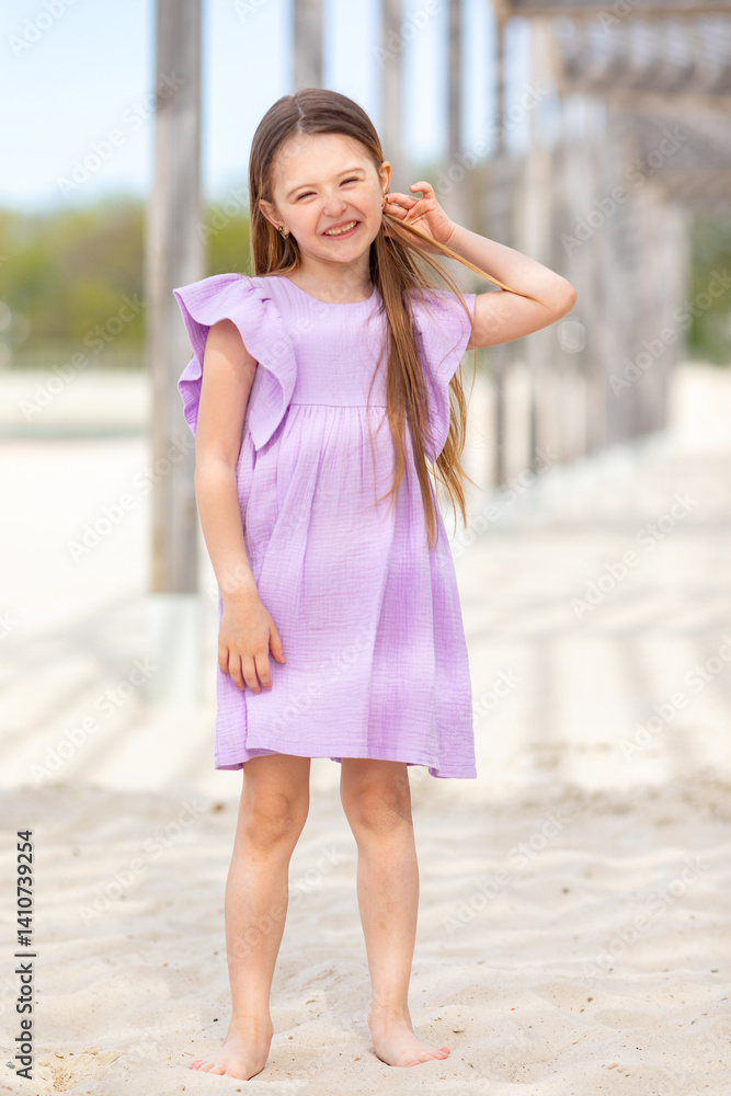 Carefree summer. Little girl in lilac dress has fun and posing on sandy beach under wooden gazebo on sunny day. Joyful childhood adventures and tropical vacation concept. Space for text.
