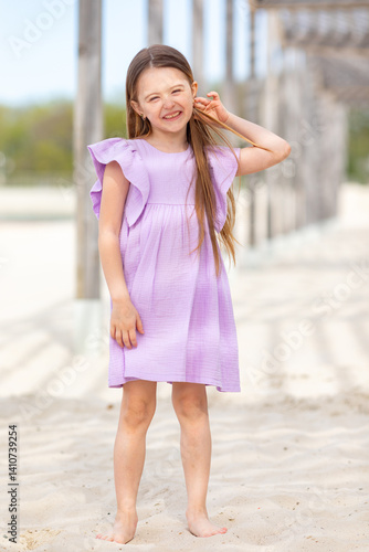 Obraz na płótnie Carefree summer. Little girl in lilac dress has fun and posing on sandy beach under wooden gazebo on sunny day. Joyful childhood adventures and tropical vacation concept. Space for text. w salonie