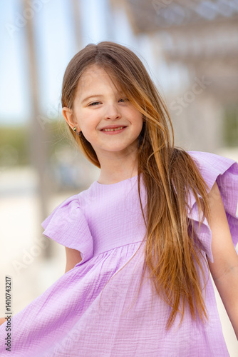 Obraz na płótnie Carefree summer. Little girl in lilac dress has fun and posing on sandy beach under wooden gazebo on sunny day. Joyful childhood adventures and tropical vacation concept. Space for text. w salonie