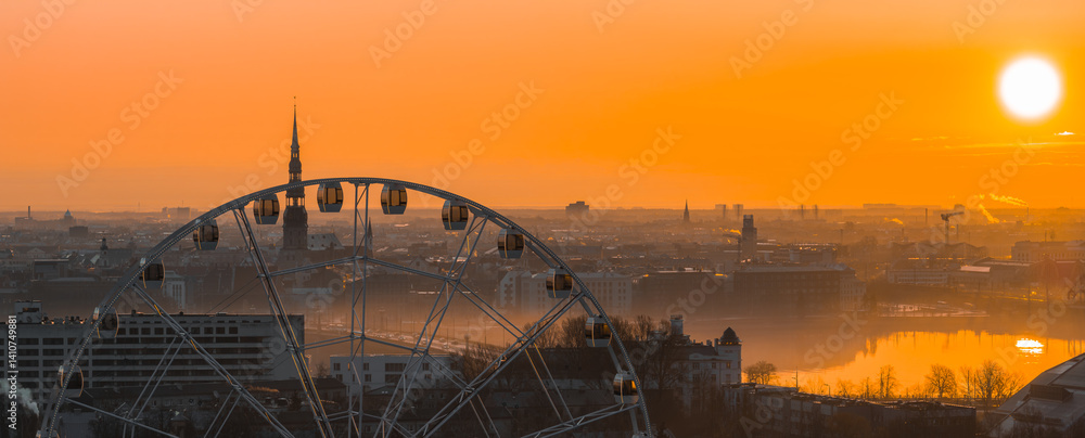 A warm sunrise illuminates Riga, Latvia, with a Ferris wheel in the foreground, St. Peter's Church spire, historic buildings, and the Daugava River.
