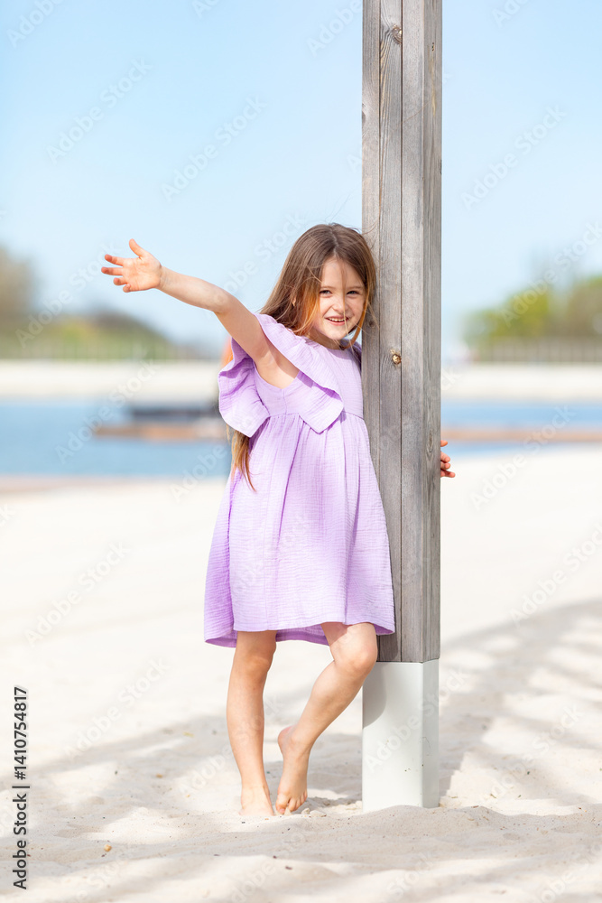 Carefree summer. Little girl in lilac dress has fun and posing on sandy beach under wooden gazebo on sunny day. Joyful childhood adventures and tropical vacation concept. Space for text.