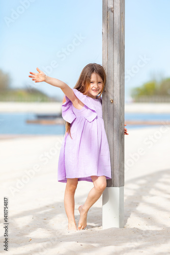 Obraz na płótnie Carefree summer. Little girl in lilac dress has fun and posing on sandy beach under wooden gazebo on sunny day. Joyful childhood adventures and tropical vacation concept. Space for text. w salonie