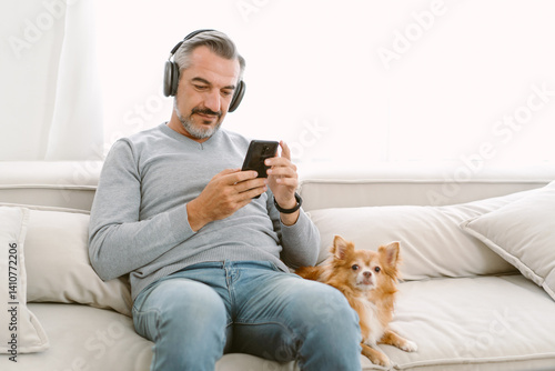Happy Middle age Caucasian man wearing headphone listening music using phone while sitting on sofa with his lovely chihuahua dog sit besides. Technology, leisure and people and Friendly Concept