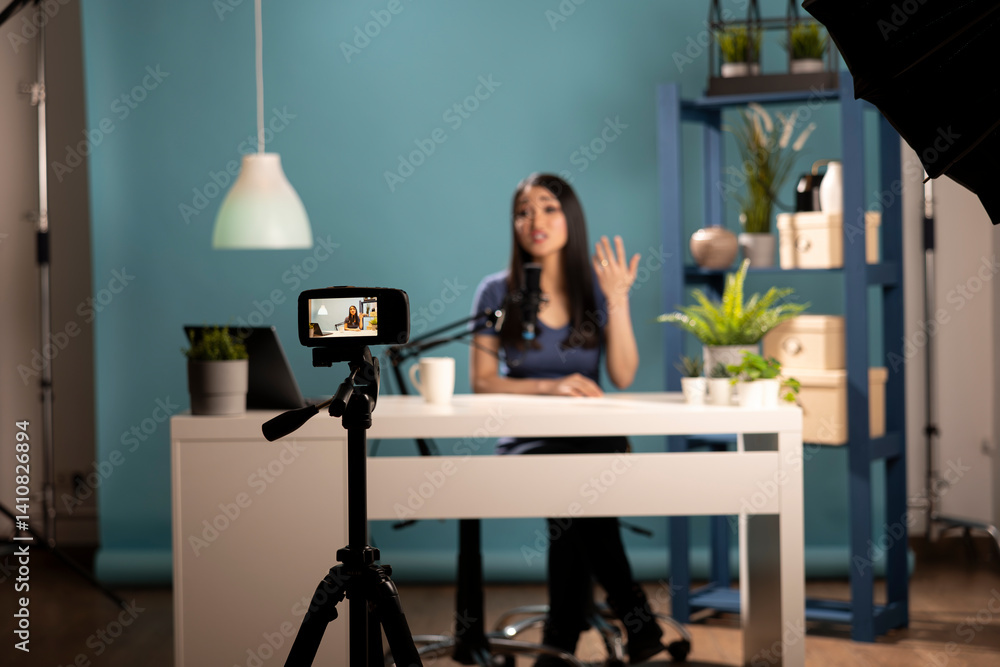 Social media influencer creating a review video in a professional studio. Asian female blogger talking to the camera, seated at a desk, using a tripod, lighting equipment, and a blue backdrop. Social media influencer creating a review video in a professional studio. Asian female blogger talking to the camera, seated at a desk, using a tripod, lighting equipment, and a blue backdrop.