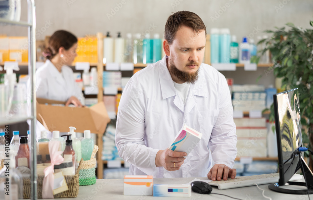 Man pharmacist uses computer at workplace. Employee reads regulations and laws, makes report, reads information about drugs. Girl assistant lays out pills merchandise in background Man pharmacist uses computer at workplace. Employee reads regulations and laws, makes report, reads information about drugs. Girl assistant lays out pills merchandise in background