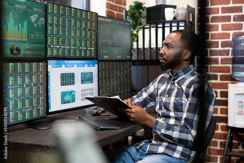 Foto Male stock market trader with a clipboard in hand, analyzing real time data on a multi monitor setup in a dimly lit office