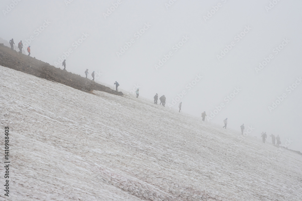 Foggy mountain landscape. Hikers go down the snow-covered slope of the volcano. Low clouds, fog and poor visibility. Kamchatka Territory, Russia. Travel, tourism and hiking on the Kamchatka Peninsula.