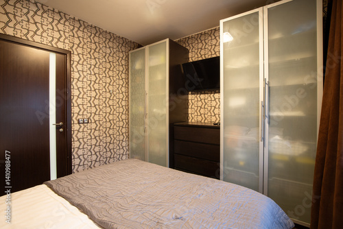Apartment interior. Bedroom. There are two large linen closets near the wall. In the niche between the wardrobes there is a chest of drawers and a black TV. The walls are covered with wallpaper.