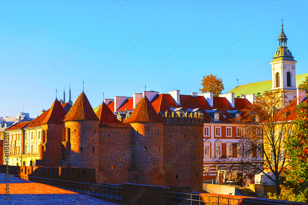 Warsaw Old Town and turrets. Medieval brick fortress with conical towers, surrounded by residential buildings with red roofs. Background with church spire rises against sky, adding contrast to scene