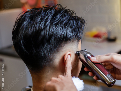 Obraz na płótnie Asian male getting a sharp undercut at barbershop, hair clipper trimming around ear with barber's guidance