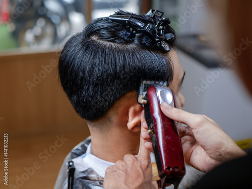 Asian man getting a haircut at a barbershop. Barber uses red clipper and wooden brush while pointing to fade line. Top hair held with black clips. Background shows mirror and warm wood decor