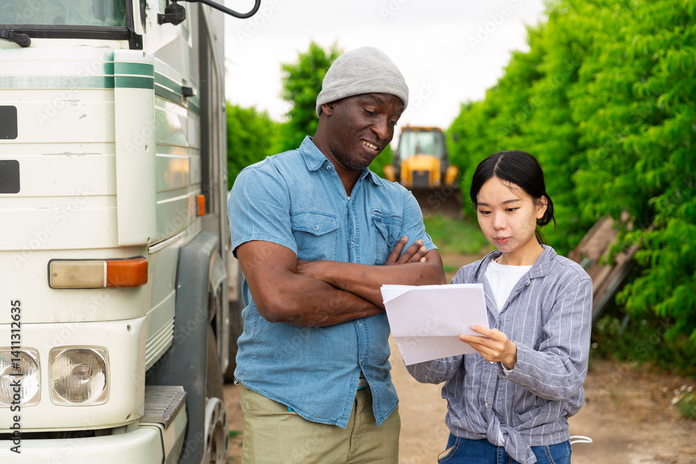 Afro american agricultural worker and asian female signing documents and talking near car at farm garden
