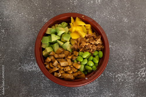 Delicious poke bowl with chicken, avocado, mango, edamame and crispy onions resting on grey table