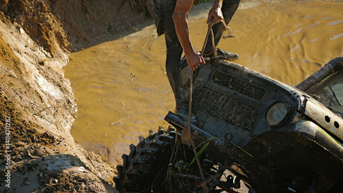 A male crew navigator stands on the hood of an SUV stuck in a muddy ditch and pulls the winch rope. Extreme racing.
