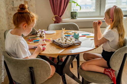 Easter preparations, red-haired and blonde girls at the table decorating Easter eggs, the girl smiles, the eggs are on a plate and on the table, the girl holds eggs in her hands, there are porcelain E
