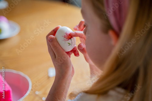 Easter preparations, a child blonde girl at the table decorates Easter eggs, eggs lie on a plate and on the table, the girl holds eggs in her hands, porcelain Easter attributes stand
