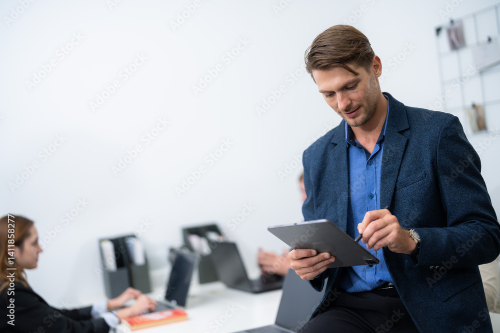 Professional man using a tablet in a modern office setting during a busy workday with colleagues nearby Professional man using a tablet in a modern office setting during a busy workday with colleagues nearby