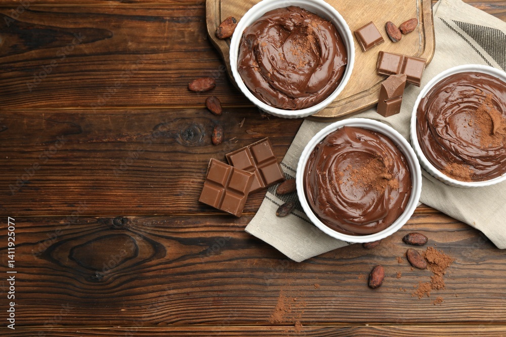 Tasty pudding in bowls, pieces of chocolate and cocoa beans on wooden table, flat lay. Space for text