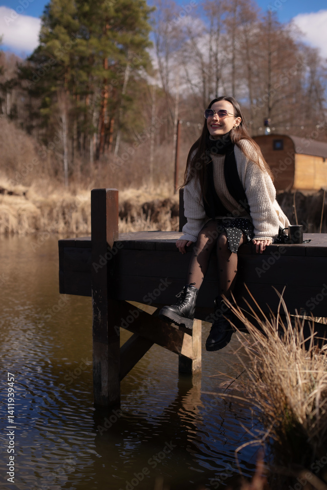 Young woman sits on a dock over a peaceful body of water, smiling and enjoying the outdoors.She wears a beige sweater, black scarf, polka-dot skirt, and boots.The background includes trees and a cabin