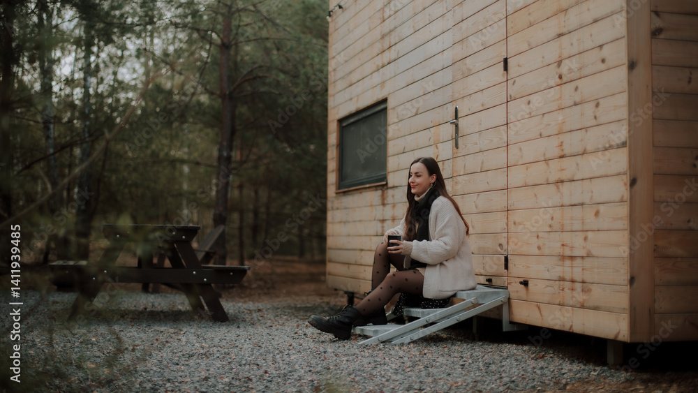 A young woman sitting on metal steps outside a wooden cabin in the forest, holding coffee mug, dressed in a cozy sweater, black scarf, and polka dot tights. A picnic table is visible in the background