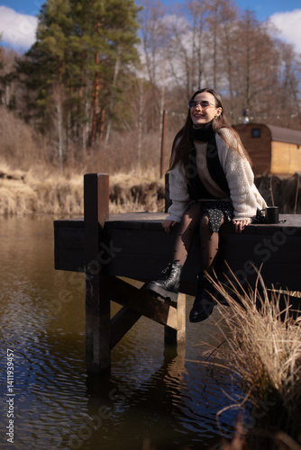 Obraz na płótnie Young woman sits on a dock over a peaceful body of water, smiling and enjoying the outdoors.She wears a beige sweater, black scarf, polka-dot skirt, and boots.The background includes trees and a cabin w salonie