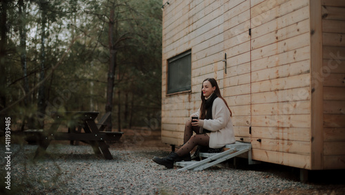 Obraz na płótnie A young woman sitting on metal steps outside a wooden cabin in the forest, holding coffee mug, dressed in a cozy sweater, black scarf, and polka dot tights. A picnic table is visible in the background w salonie