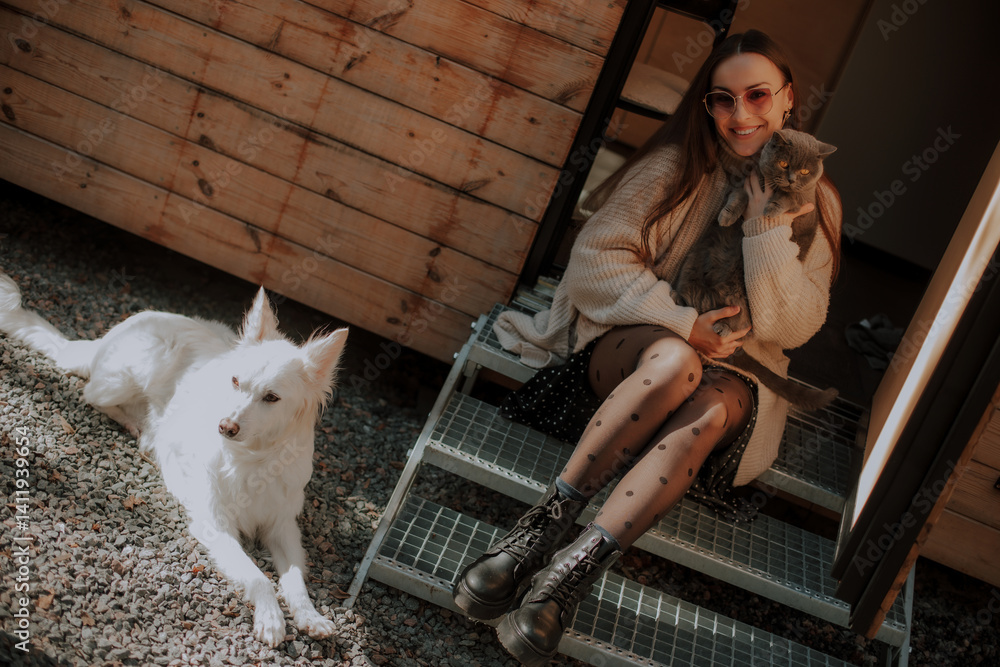 A woman sits on outdoor steps, smiling while holding a gray cat in her arms. A white dog lies next to her on the gravel, with a cozy wooden cabin in the background. Casual, relaxed atmosphere.