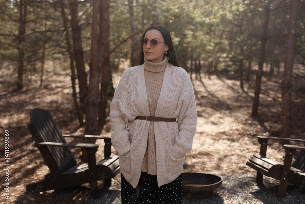 A woman in a cozy sweater and scarf stands in the woods, wearing sunglasses and a belt. She poses confidently near outdoor seating, with soft sunlight filtering through the trees.