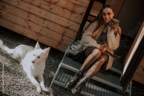 Obraz na płótnie A woman sits on outdoor steps, smiling while holding a gray cat in her arms. A white dog lies next to her on the gravel, with a cozy wooden cabin in the background. Casual, relaxed atmosphere. w salonie