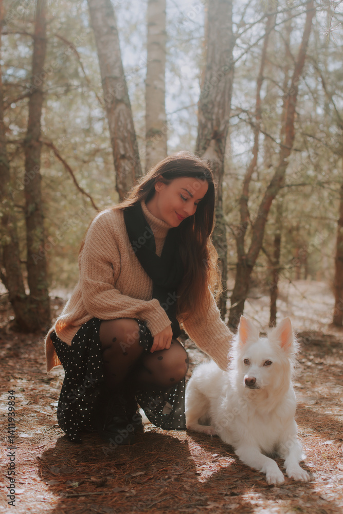 A woman in a cozy beige sweater and black skirt kneels in the forest, gently petting her white dog. Sunlight filters through the trees, creating a warm, peaceful moment in nature.