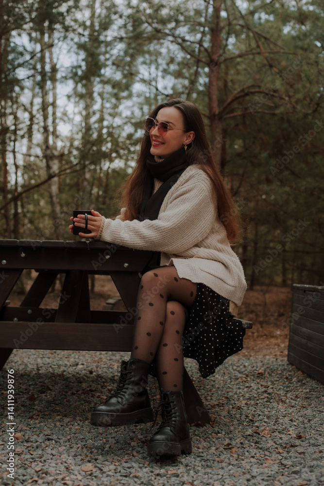 A woman sitting at a picnic table in the forest, holding a black cup and smiling. She wears a cozy sweater, black tights with polka dots, and boots, enjoying a serene moment in nature.