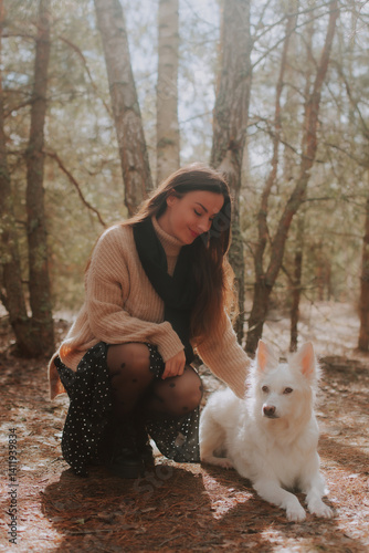 Obraz na płótnie A woman in a cozy beige sweater and black skirt kneels in the forest, gently petting her white dog. Sunlight filters through the trees, creating a warm, peaceful moment in nature. w salonie