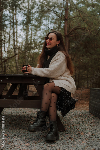 Obraz na płótnie A woman sitting at a picnic table in the forest, holding a black cup and smiling. She wears a cozy sweater, black tights with polka dots, and boots, enjoying a serene moment in nature. w salonie