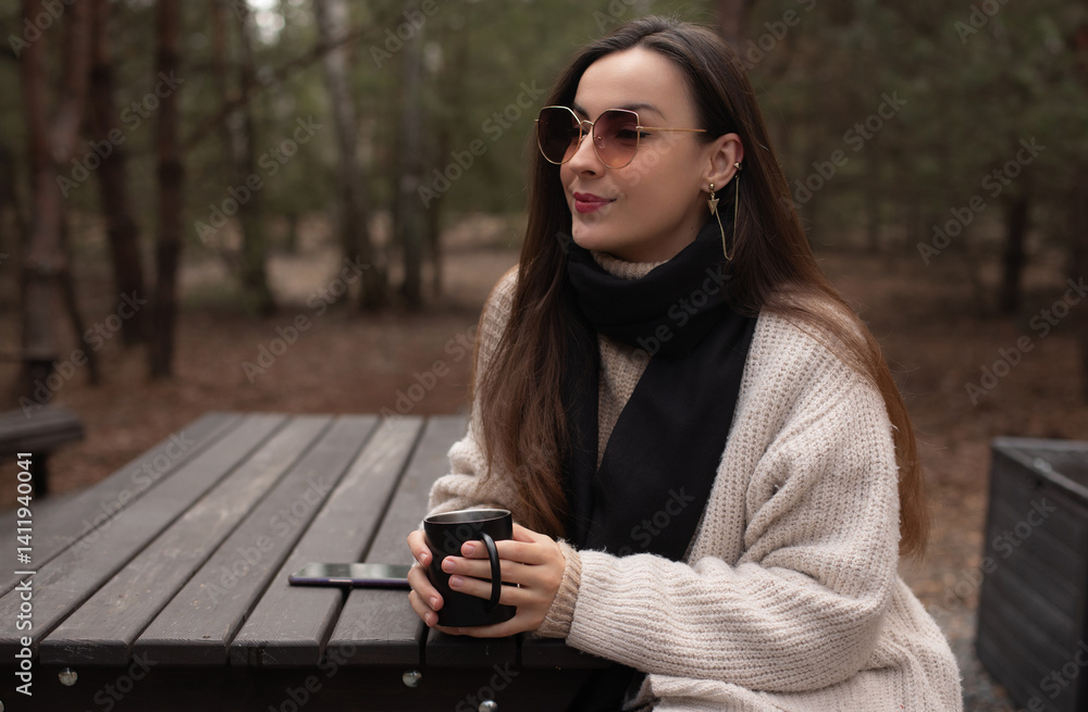 A woman sitting at an outdoor table in the forest, wearing a cozy sweater and black scarf, holding a cup of coffee. She wears sunglasses and enjoys a peaceful, serene moment in nature.