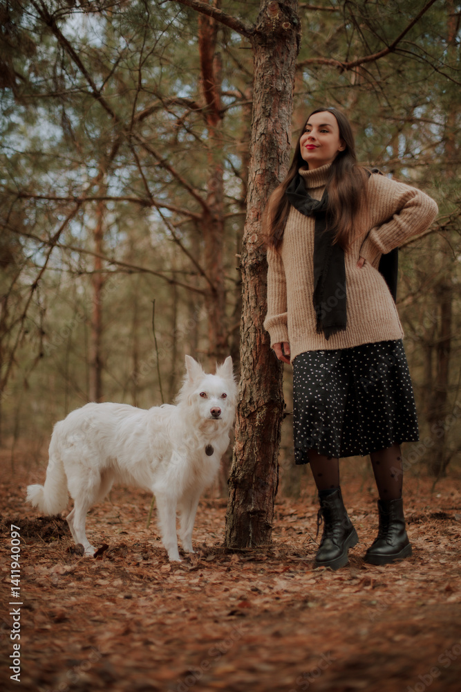 A woman in a cozy beige sweater and black skirt stands in the forest with her white dog, both posing naturally by a tree. The scene is serene, with autumn leaves on the ground and a calm atmosphere.