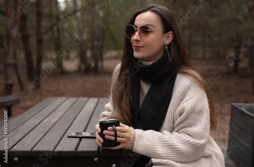 Obraz na płótnie A woman sitting at an outdoor table in the forest, wearing a cozy sweater and black scarf, holding a cup of coffee. She wears sunglasses and enjoys a peaceful, serene moment in nature. w salonie