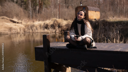 A woman sitting on a wooden dock by a peaceful pond, enjoying a cup of coffee. She smiles, wearing a cozy sweater, black tights with polka dots, and sunglasses, surrounded by natural beauty.
