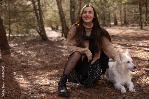 Obraz na płótnie A woman in a cozy sweater and polka dot skirt kneels in the forest beside her white dog, smiling and gently petting it. The natural setting creates a serene and joyful moment. w salonie