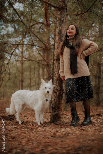 Obraz na płótnie A woman in a cozy beige sweater and black skirt stands in the forest with her white dog, both posing naturally by a tree. The scene is serene, with autumn leaves on the ground and a calm atmosphere. w salonie