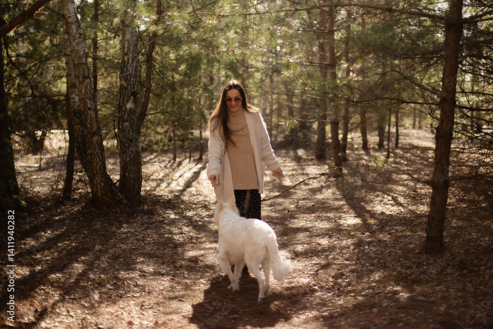 A woman in a cozy sweater and cardigan walks through the forest, playfully interacting with her white dog. Sunlight filters through the trees, creating a warm, peaceful outdoor scene.