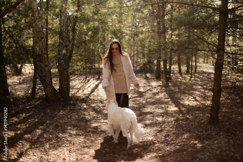 Obraz na płótnie A woman in a cozy sweater and cardigan walks through the forest, playfully interacting with her white dog. Sunlight filters through the trees, creating a warm, peaceful outdoor scene. w salonie
