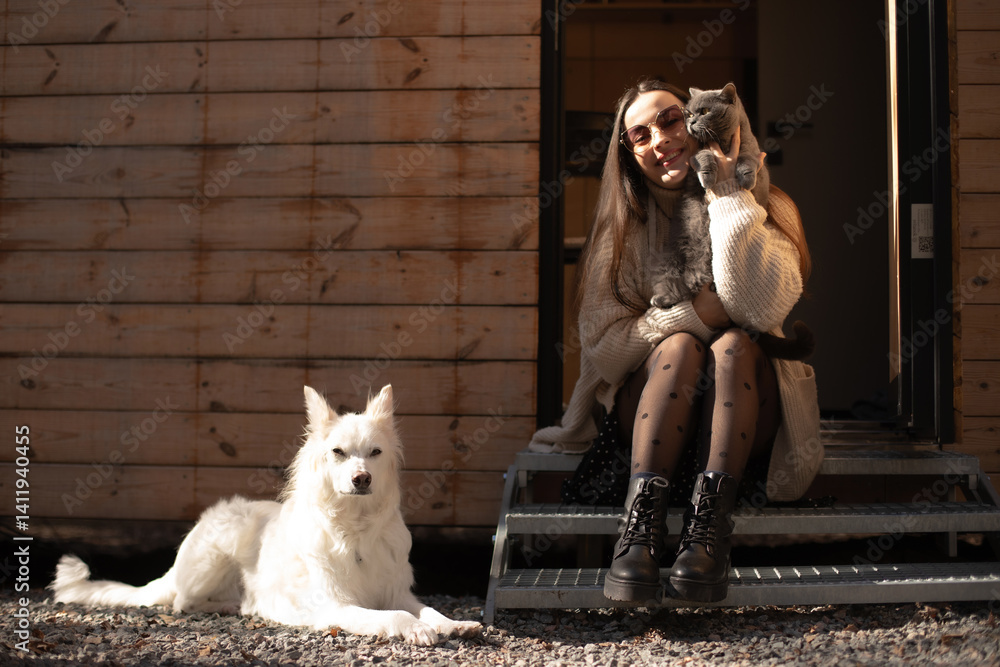 A woman sitting on steps outside a wooden cabin, holding a cat and smiling. A white dog rests nearby. The scene is sunlit, showcasing a peaceful moment with pets in a natural setting.