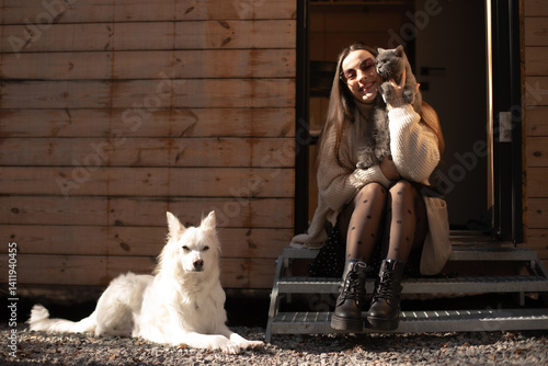 Obraz na płótnie A woman sitting on steps outside a wooden cabin, holding a cat and smiling. A white dog rests nearby. The scene is sunlit, showcasing a peaceful moment with pets in a natural setting. w salonie