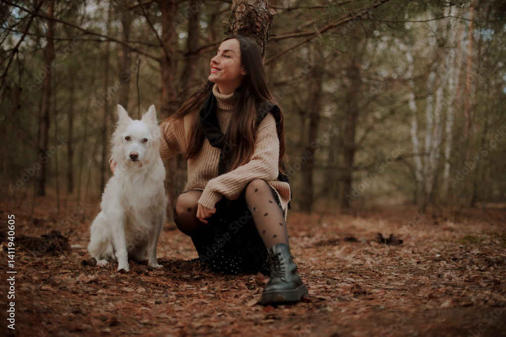A woman with long hair wearing a cozy sweater and boots, kneeling in the forest with her white dog, both surrounded by autumn leaves, smiling and enjoying the peaceful outdoors.
