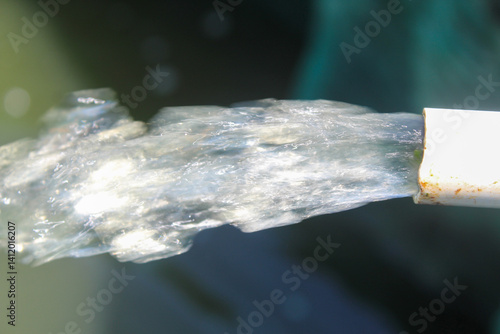 Close-up of water gushing from a white pipe, captured mid-motion with sunlight highlighting the flow. The background is dark, creating a striking contrast with the clear, dynamic water stream.