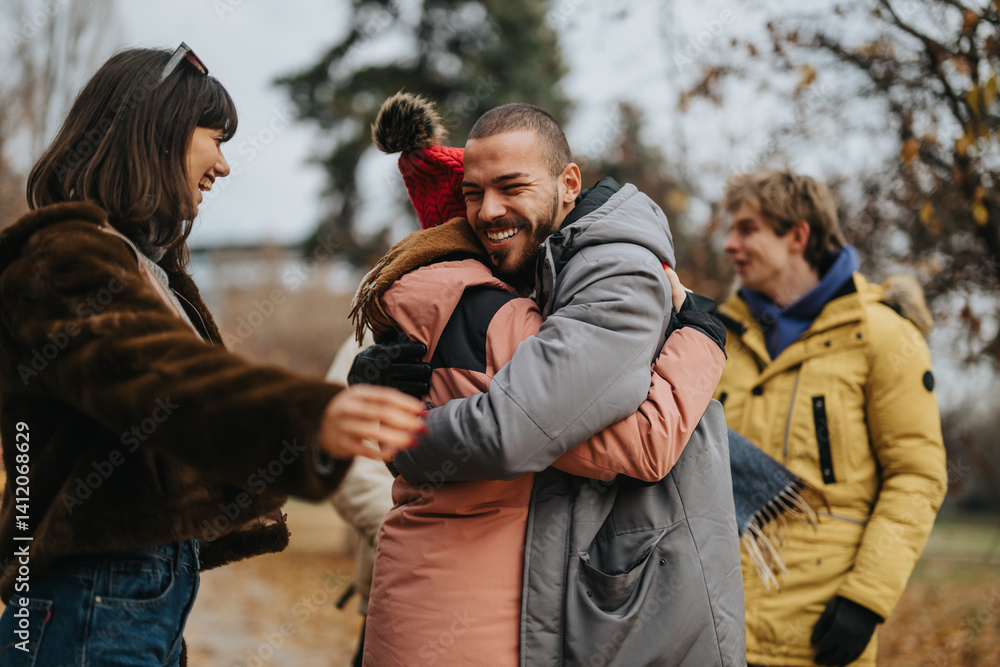 Fototapeta premium Friends gather in a park during winter, dressed in warm clothing. They share joyful hugs and smiles, surrounded by nature. The scene conveys friendship, warmth, and happy winter moments.